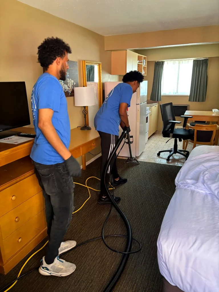 Two crew members cleaning the carpet in a guest room with a bed and desk