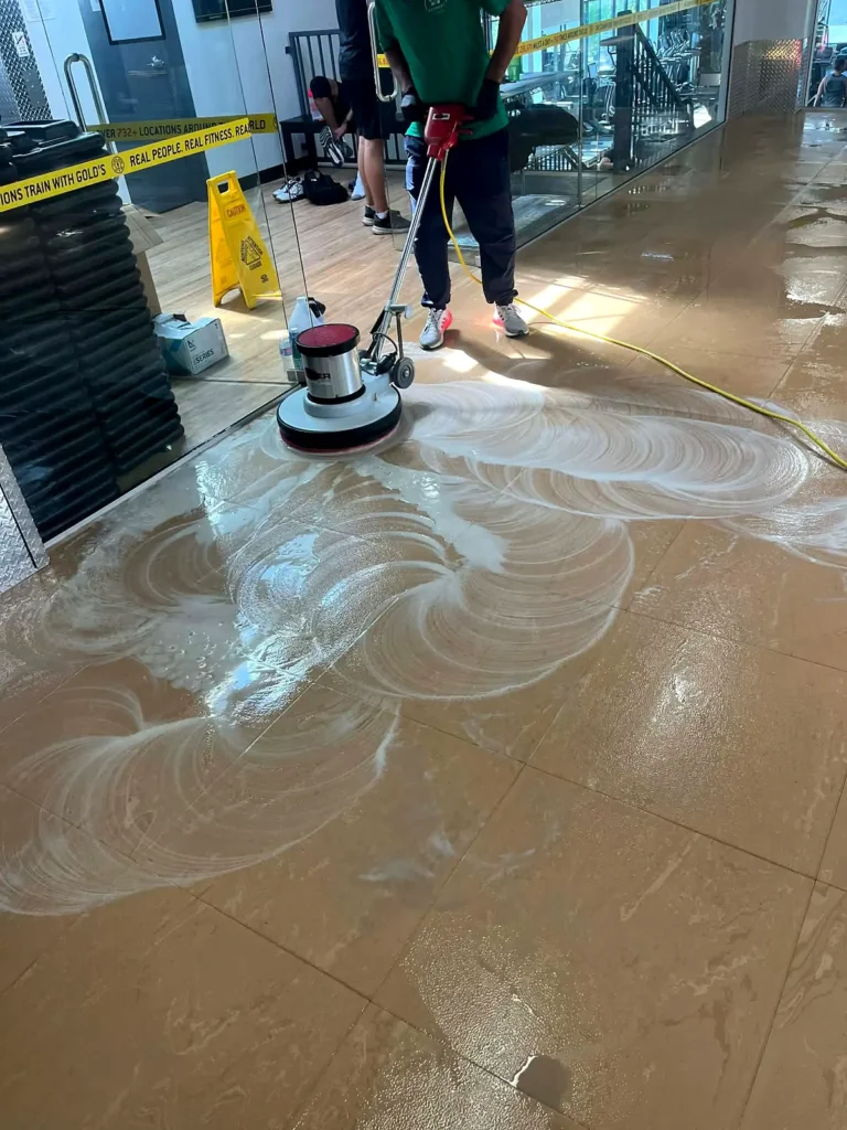 Crew member scrubbing a gym floor with a rotary machine behind a caution sign