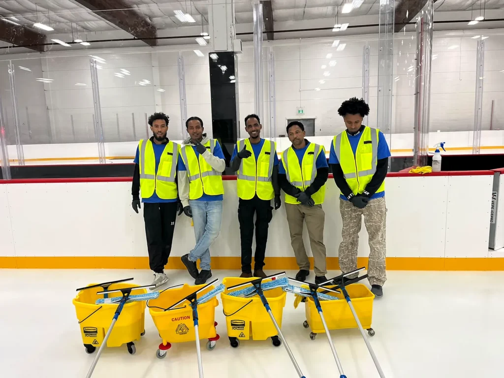 Five Everest crew members in hi-vis vests with mop buckets and squeegees beside the rink boards