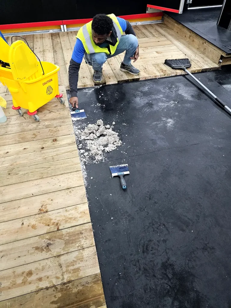 Crew member scraping construction residue off a black rubber floor beside the rink boards