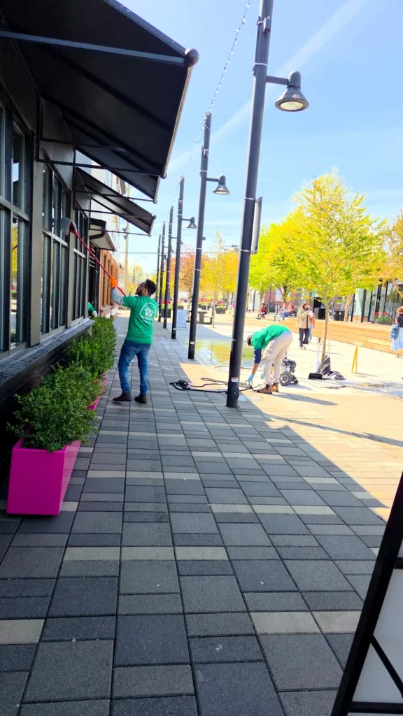 Crew cleaning storefront windows and sidewalk beside pink planters at a restaurant
