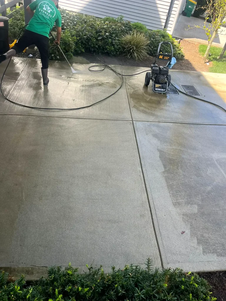 Crew member pressure washing a concrete driveway, cleaned and uncleaned areas visible